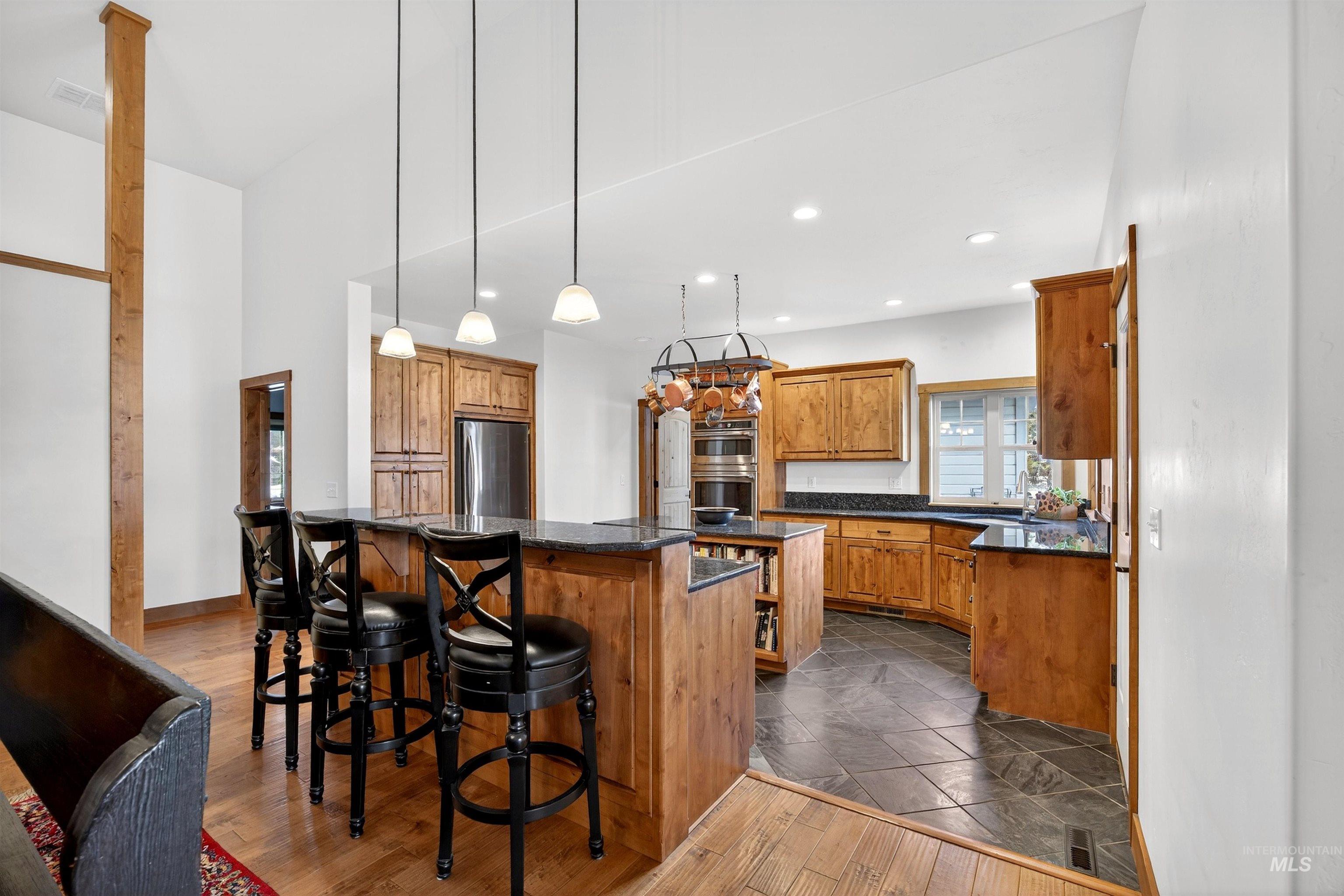 Spacious kitchen featuring rich wood cabinetry, dark countertops, a breakfast bar with pendant lighting, stainless steel appliances, and slate tile flooring