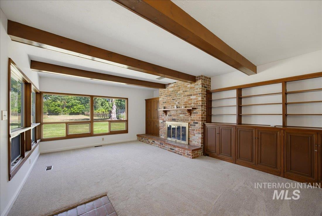 Spacious room featuring exposed wood ceiling beams, extensive window framing, a brick fireplace with a hearth, and built-in wood shelving with lower cabinetry