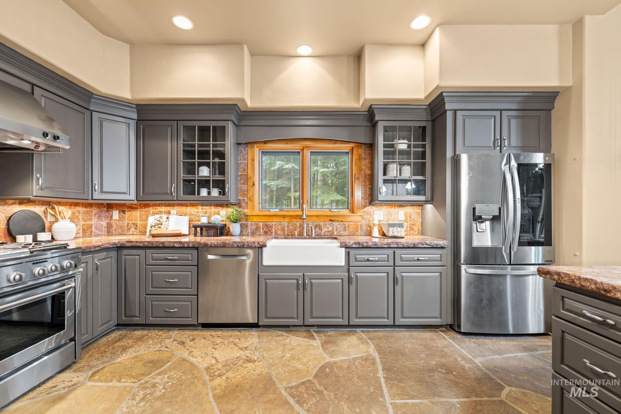Kitchen with gray cabinetry, glass insert cabinets, stainless steel appliances, backsplash, and stone tile floors