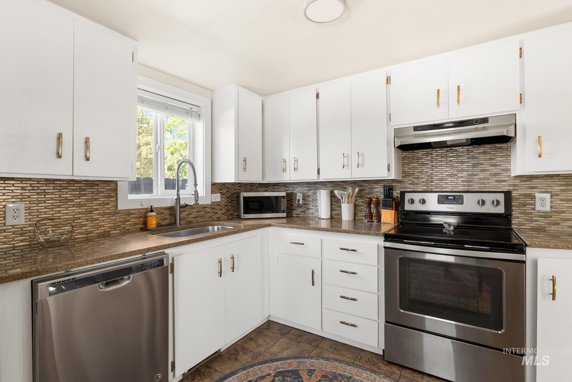 Kitchen featuring white cabinetry with gold-finish hardware, stainless steel appliances, mosaic tile backsplash, and a window above the sink