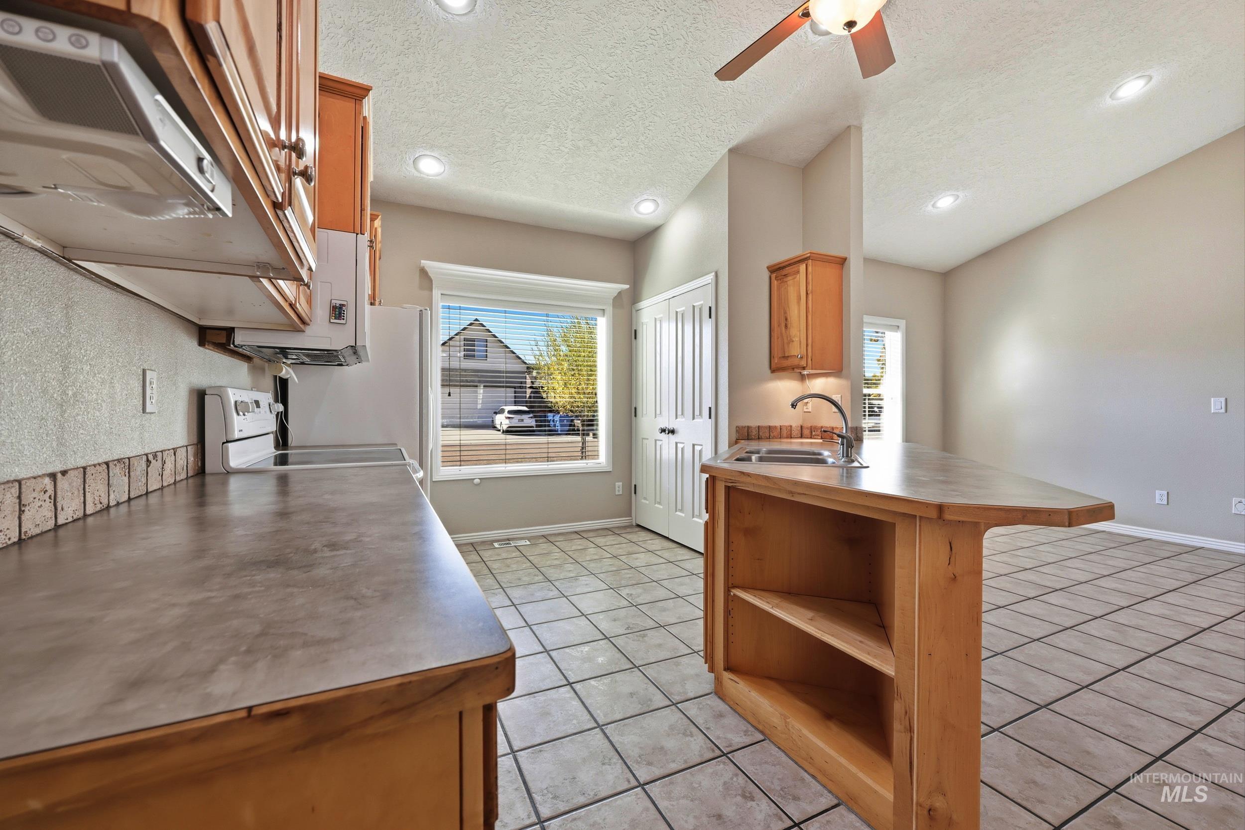 Kitchen featuring extractor fan, a textured ceiling, light tile patterned floors, brown cabinetry, and healthy amount of natural light
