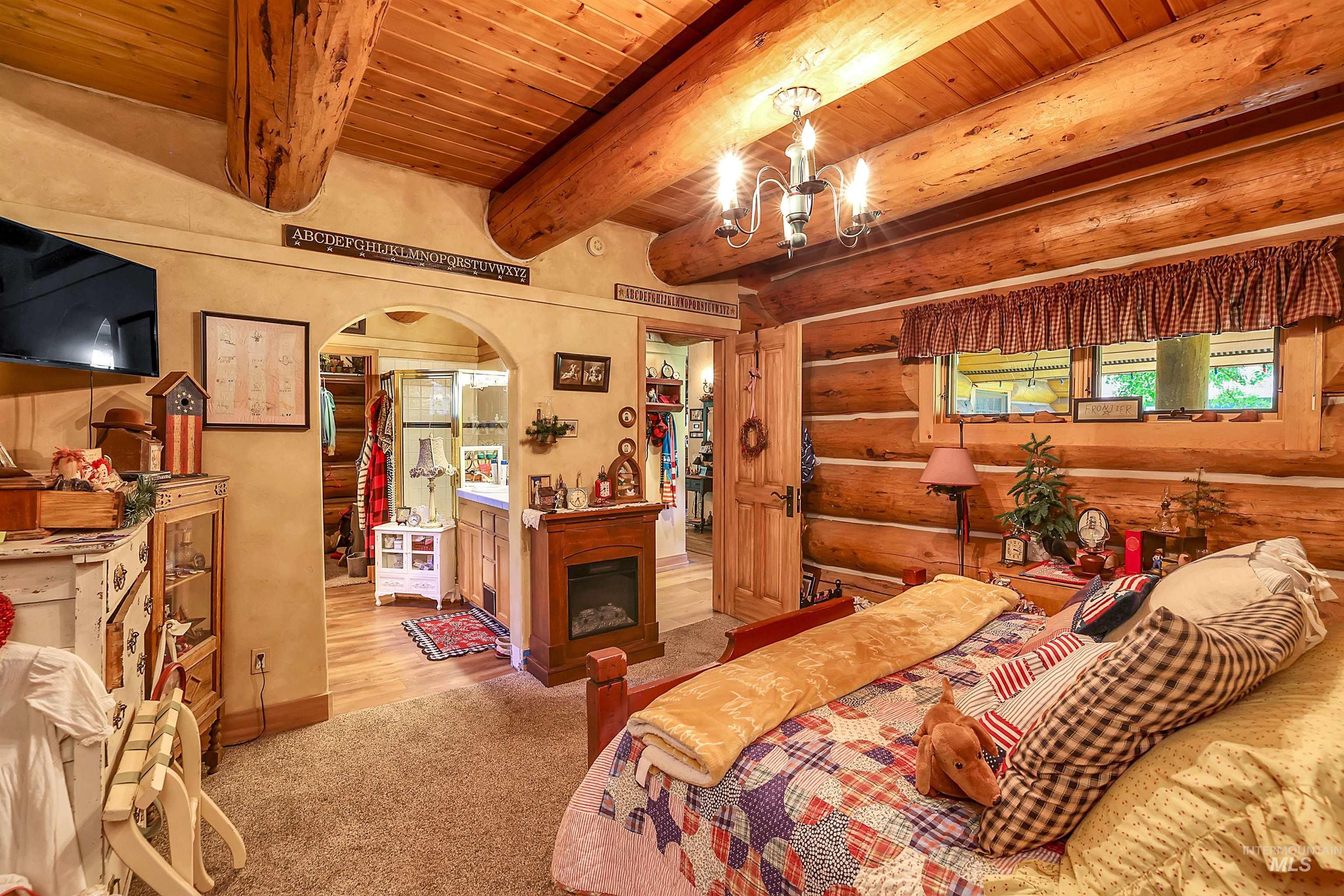 Carpeted bedroom with arched walkways, log walls, a wood ceiling with exposed beams, a chandelier, and a fireplace