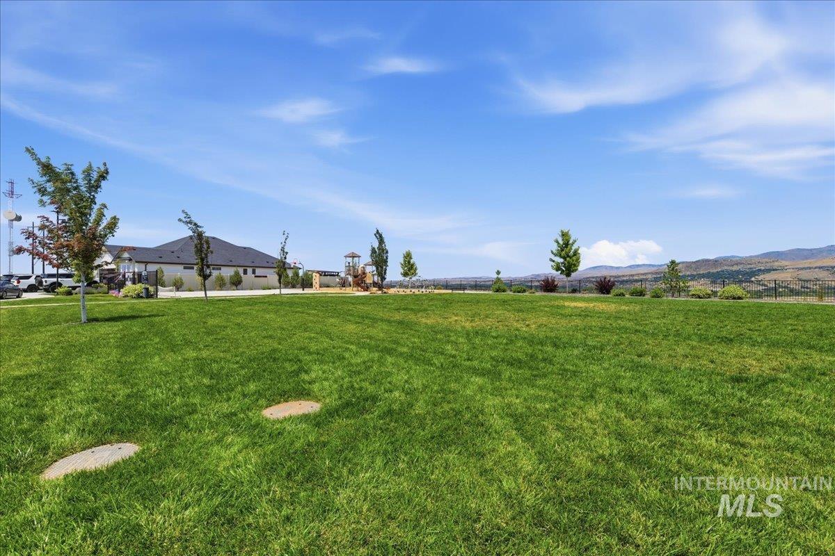 Expansive grass area with a distant structure featuring a dark roof and a playset, surrounded by established trees and landscaping