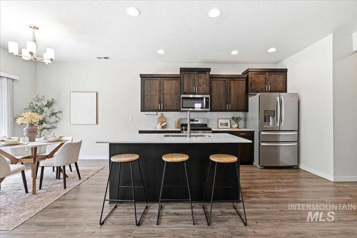 Open-concept kitchen and dining area featuring wood-finish flooring, recessed lighting, dark wood cabinetry, a center island with a light-colored countertop and integrated sink, and stainless steel appliances