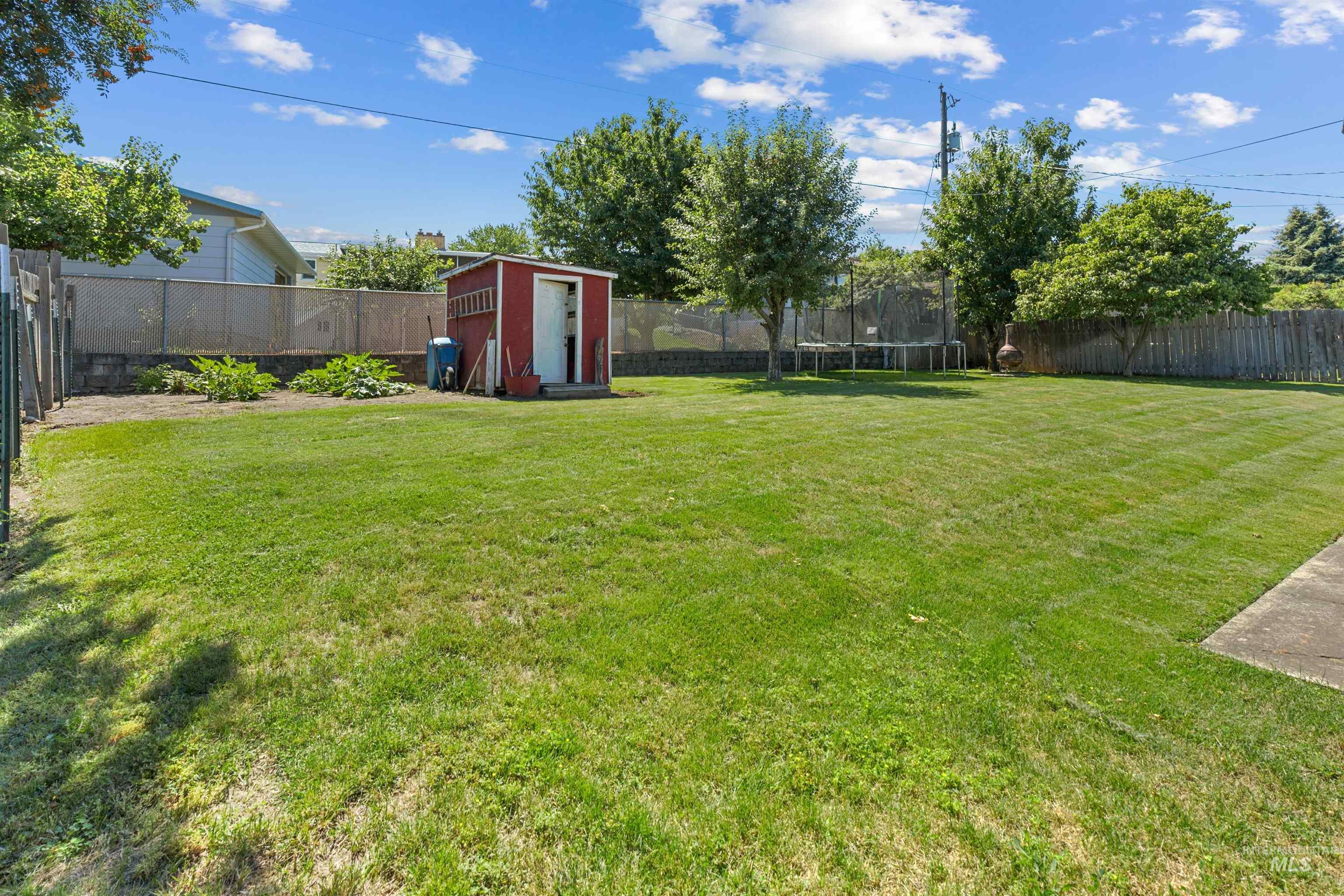 Expansive lawn area featuring a red storage shed, concrete patio, mature trees, and a combination of wood and chain-link fencing