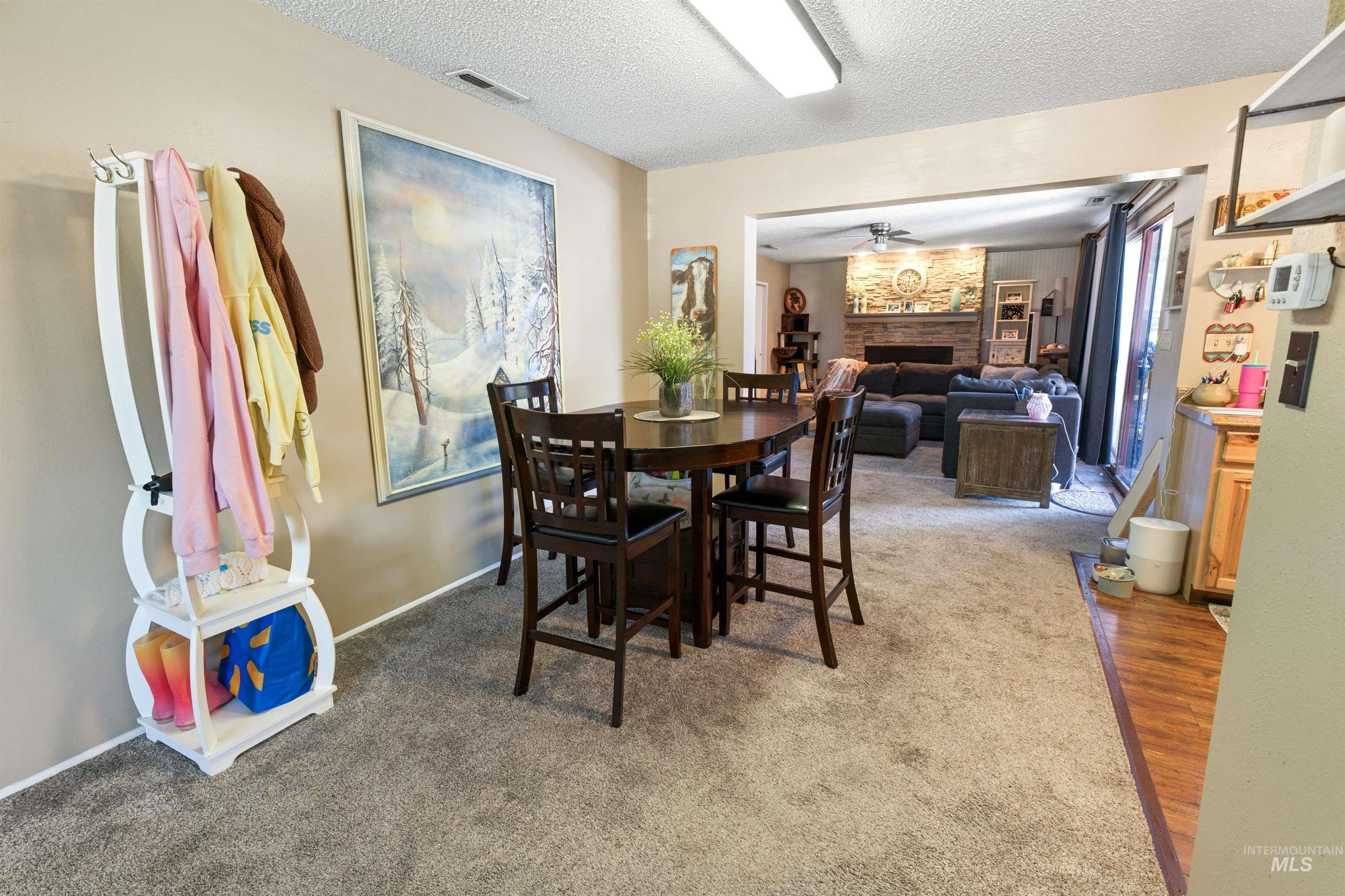 Carpeted dining area with a dark wood-finish table and chairs, featuring an opening to a carpeted living space with a stone-clad fireplace and ceiling fan