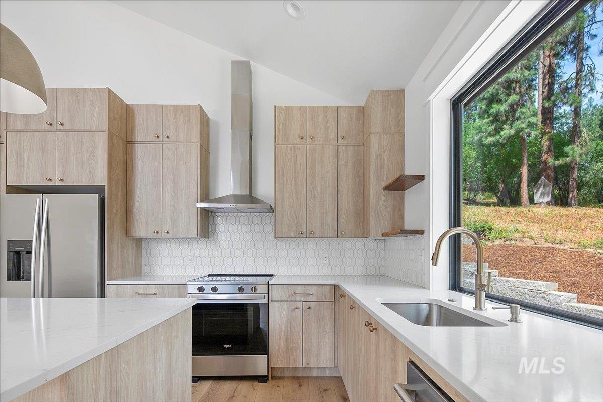 Kitchen featuring light brown cabinetry, open shelves, stainless steel appliances, vaulted ceiling, and wall chimney exhaust hood