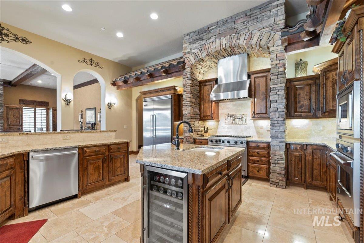 Kitchen featuring built in appliances, wine cooler, wall chimney range hood, decorative backsplash, and light stone countertops