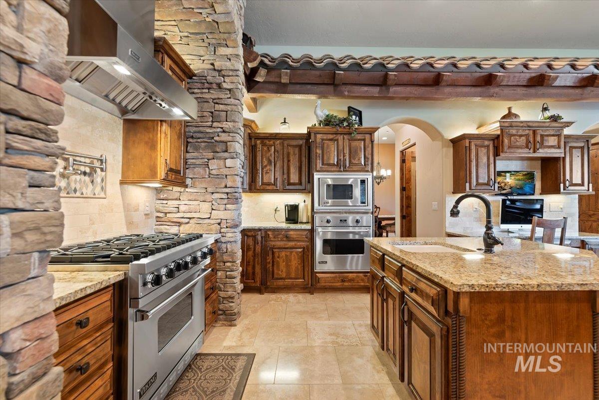 Kitchen with appliances with stainless steel finishes, exhaust hood, decorative backsplash, arched walkways, and light stone countertops