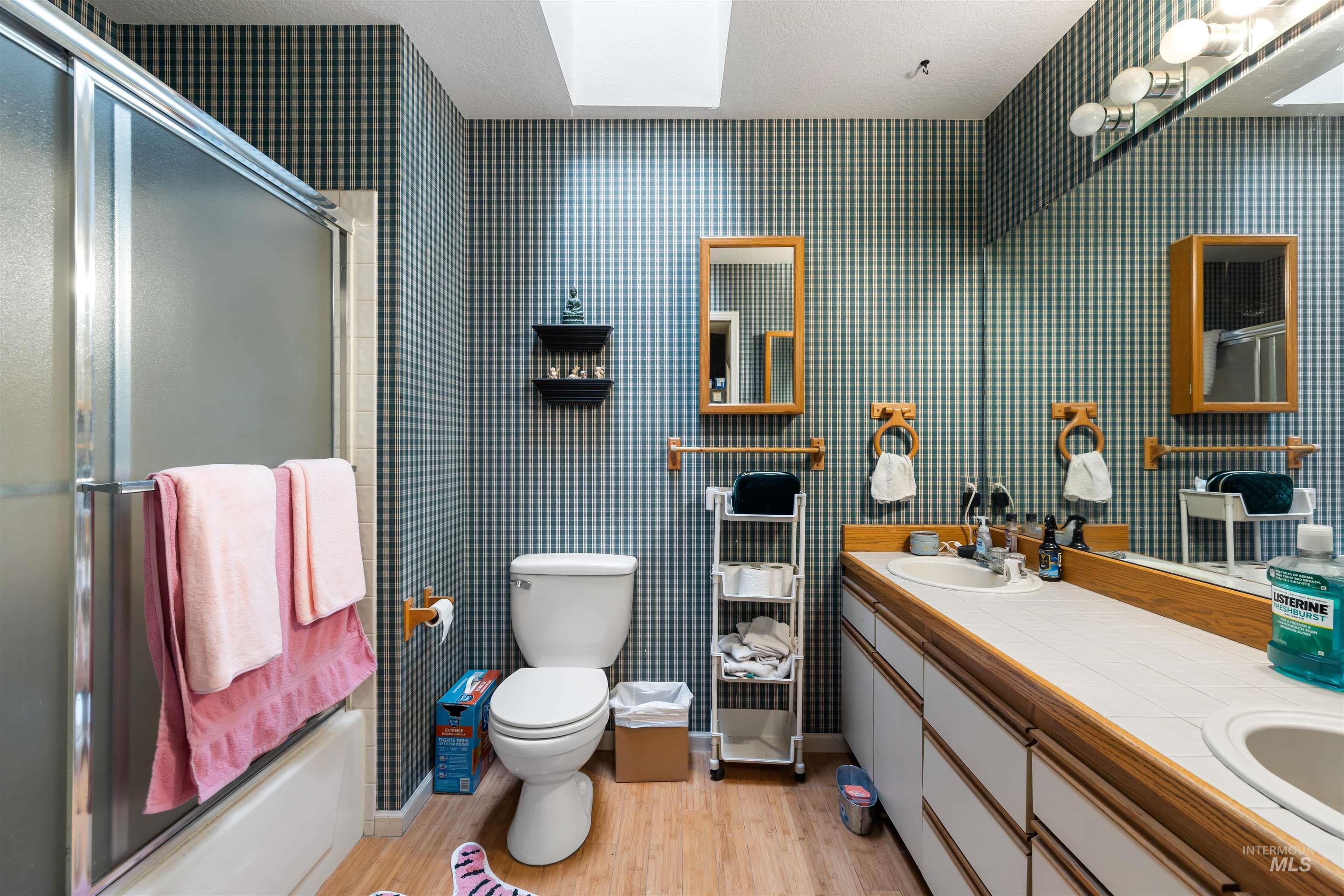 Bathroom with double vanity, light wood finished floors, shower / bath combination with glass door, and a skylight
