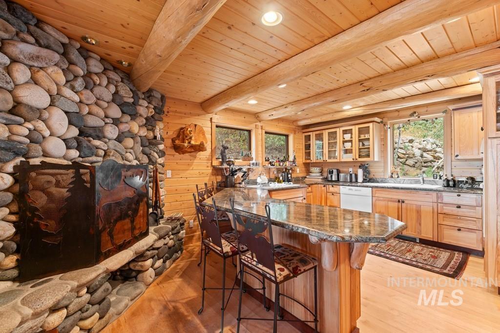 Stone fireplace with a decorative metal screen, hardwood flooring, wood plank ceilings with exposed beams, wood cabinetry with glass-front uppers, and a granite-topped kitchen island with seating