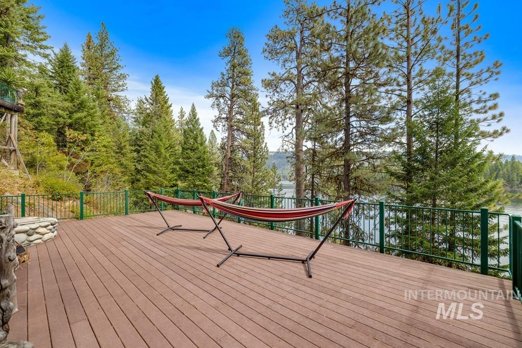 Expansive composite deck featuring a green metal railing, a built-in stone fire pit, and surrounded by mature pine trees and a body of water