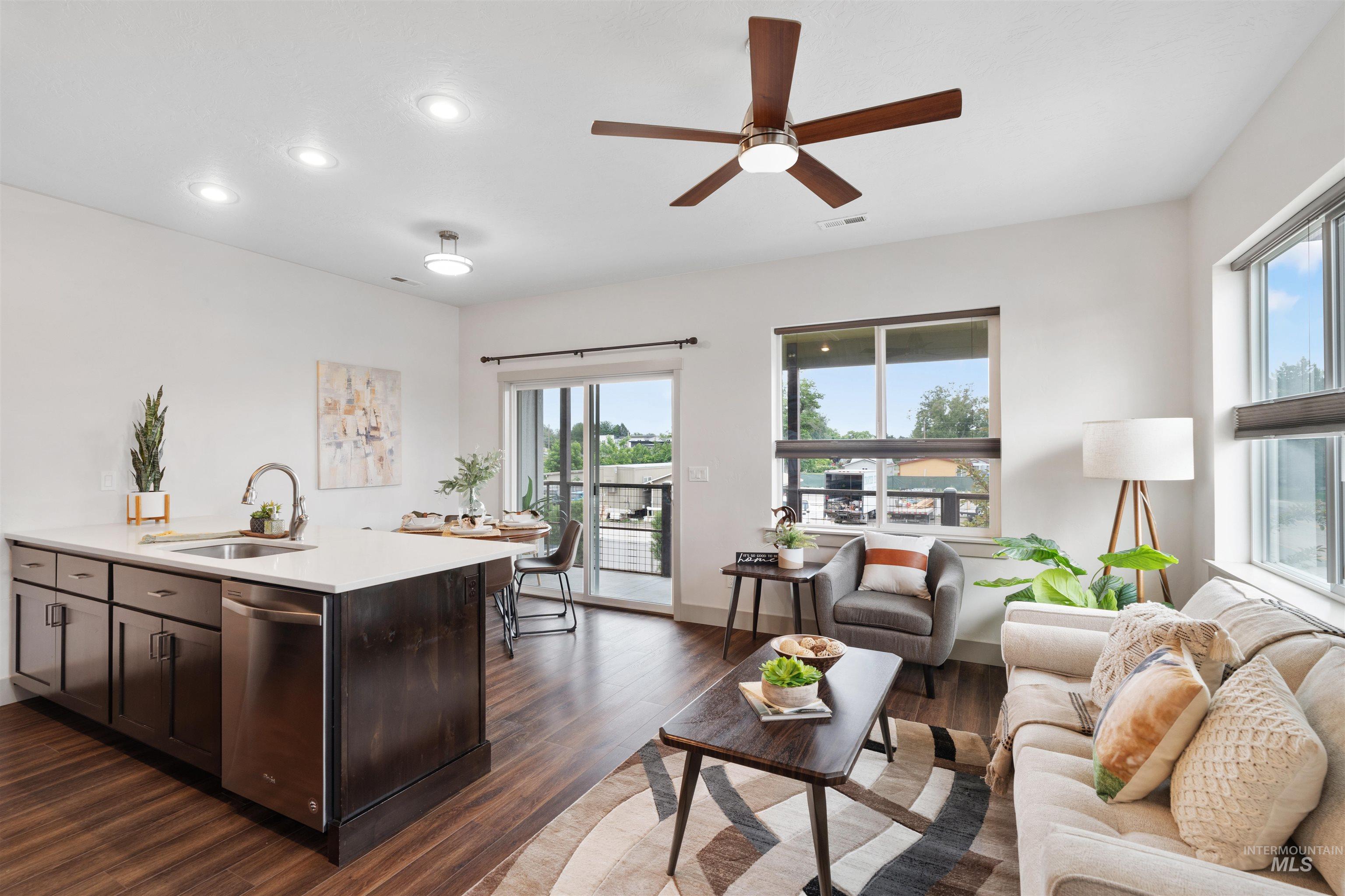 Open-concept living area featuring wood-finish flooring, a contemporary kitchen island with white countertop and stainless steel appliances, and abundant natural light from large windows and a sliding glass door
