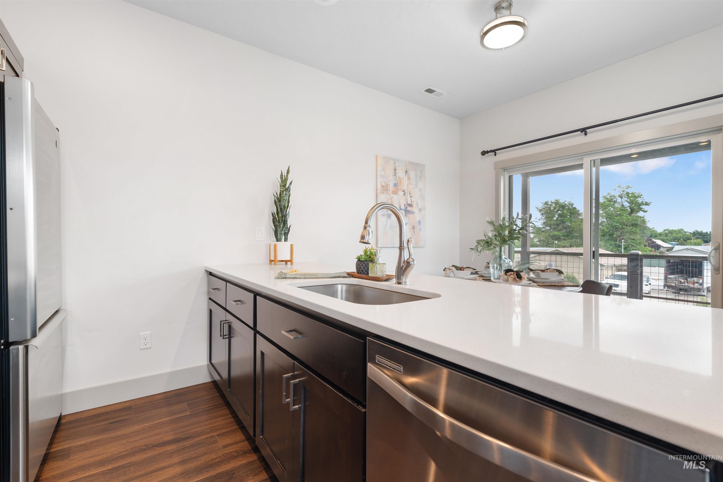 Kitchen island featuring white countertop, dark wood-finish cabinetry, stainless steel sink with gooseneck faucet, and integrated stainless steel dishwasher