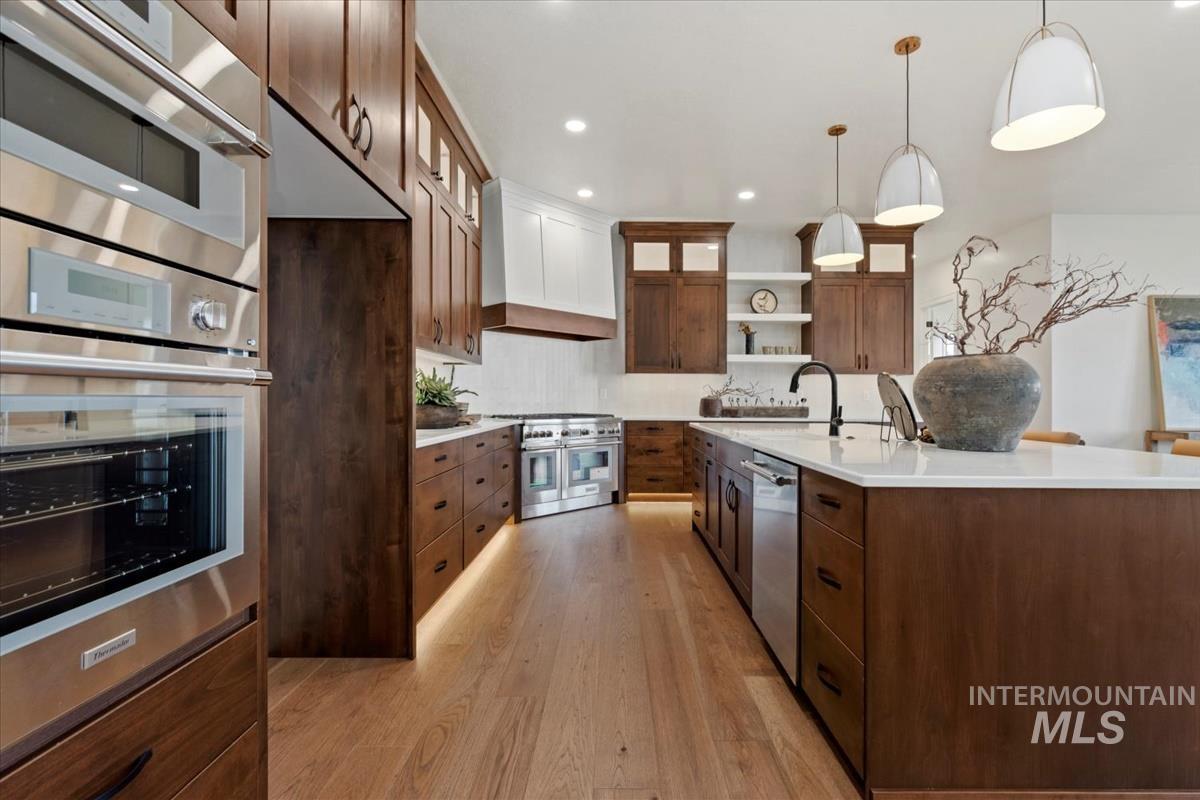 Kitchen featuring glass insert cabinets, open shelves, appliances with stainless steel finishes, decorative light fixtures, and custom exhaust hood