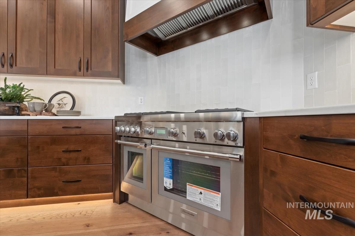 Kitchen with double oven range, custom range hood, light wood-style floors, decorative backsplash, and brown cabinets