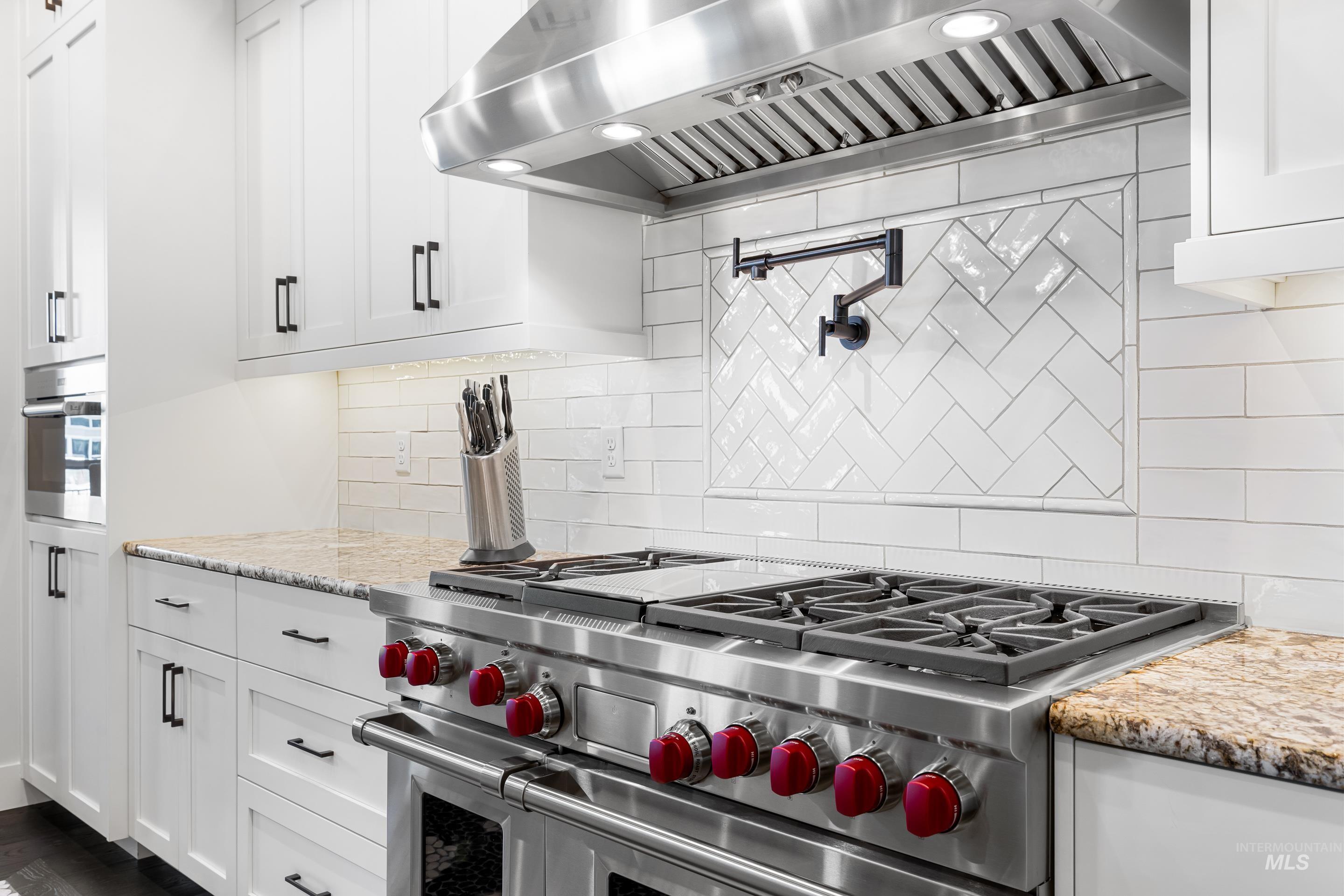 Kitchen with wall chimney range hood, backsplash, white cabinets, stainless steel appliances, and light stone counters