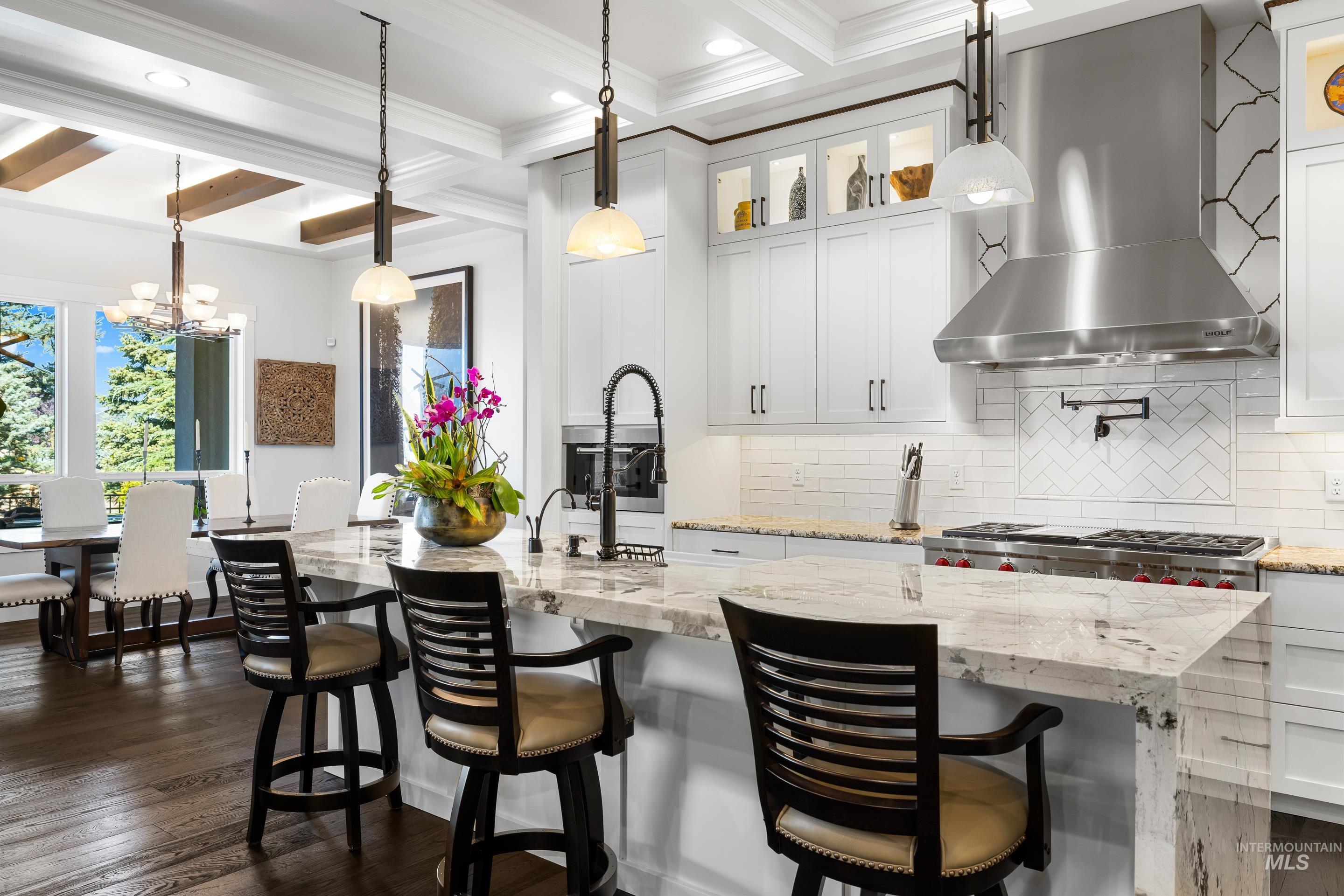 Kitchen with hanging light fixtures, wall chimney range hood, backsplash, light stone counters, and beam ceiling