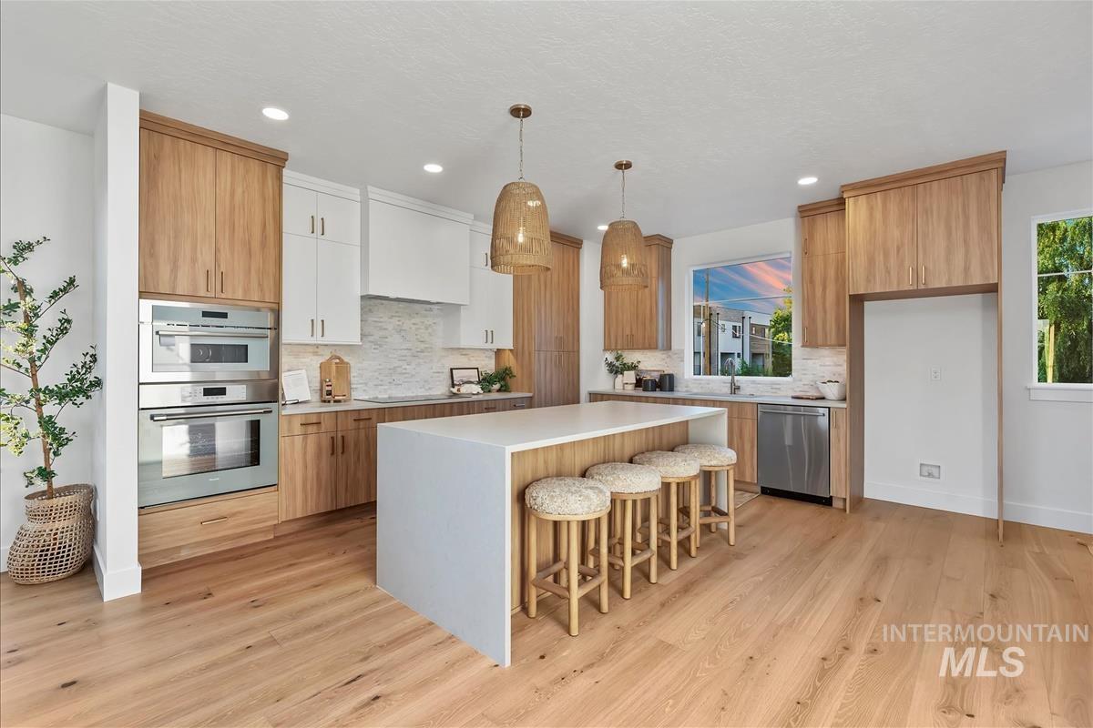 Bright kitchen featuring a waterfall island with seating, a stainless steel appliance suite including double ovens and a dishwasher, wood-finish flooring, and a patterned tile backsplash