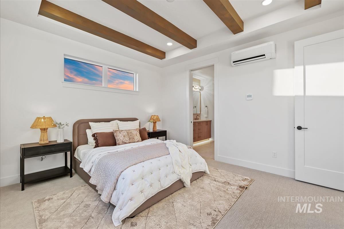 Bedroom featuring overhead wood beams, a horizontal window, wall-mounted climate control, and an open doorway leading to a bathroom with a vanity