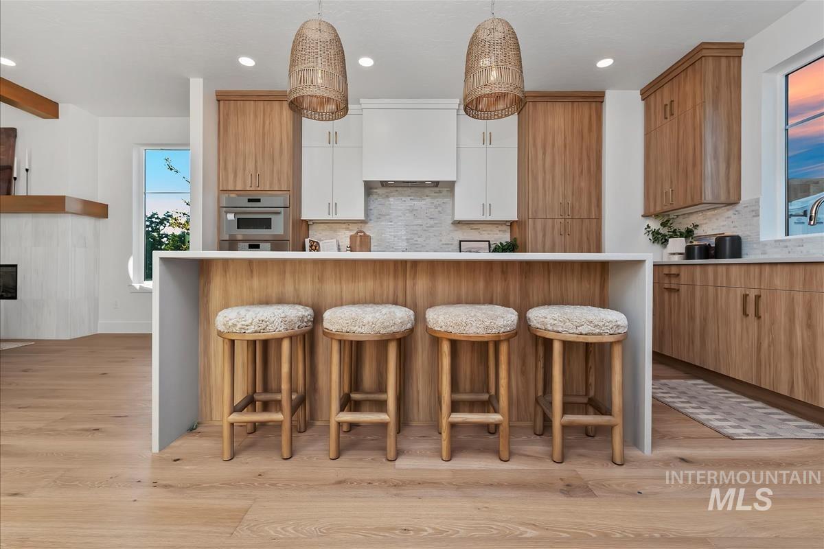 Kitchen island with white waterfall countertop, light wood-finish cabinetry, white upper cabinets, stainless steel built-in microwave, and pendant lighting