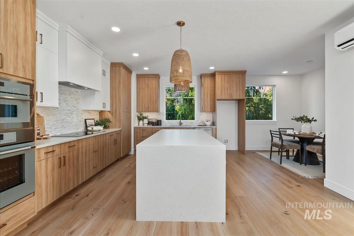 Modern kitchen featuring a waterfall island, wood-finish flooring, two-tone cabinetry with light wood and white finishes, stainless steel built-in double ovens, and a patterned tile backsplash