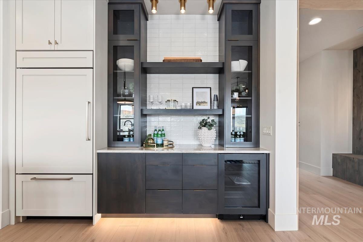 Indoor dry bar with wine cooler, paneled refrigerator, light wood-style floors, open shelves, and decorative backsplash