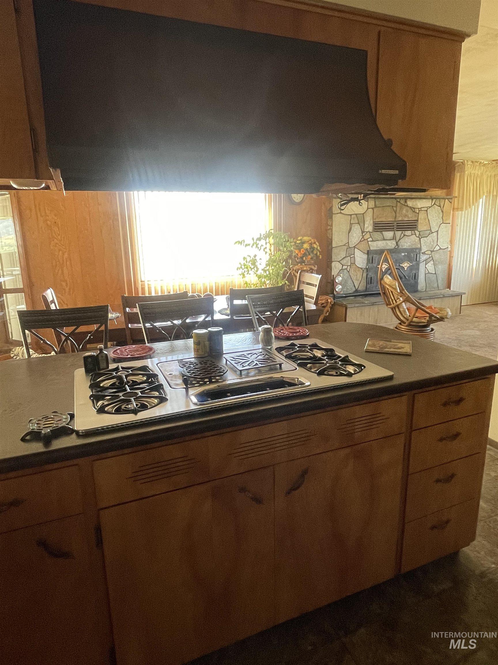 Kitchen featuring stainless steel gas stovetop, brown cabinetry, a stone fireplace, and under cabinet range hood
