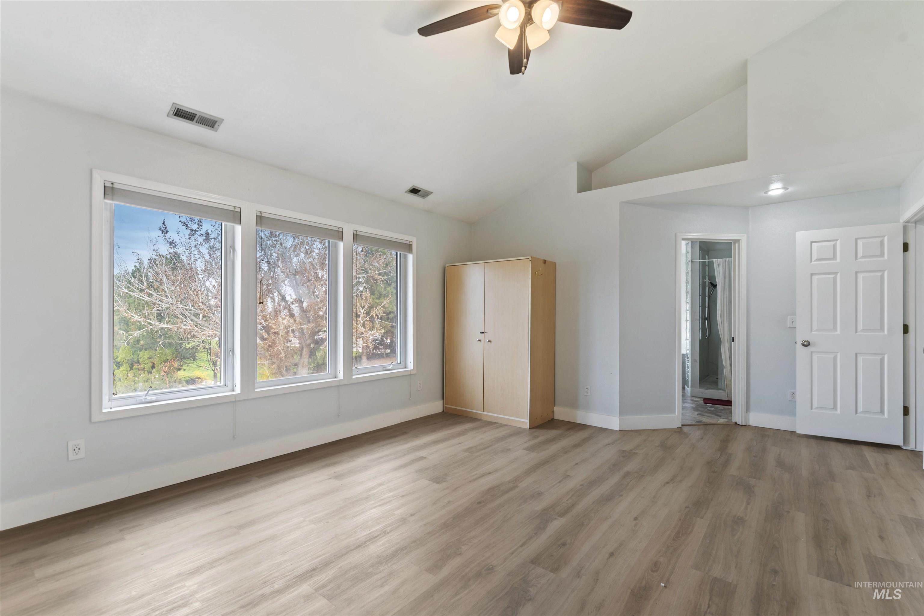 Unfurnished bedroom with light wood finished floors, multiple windows, ceiling fan, ensuite bathroom, and high vaulted ceiling