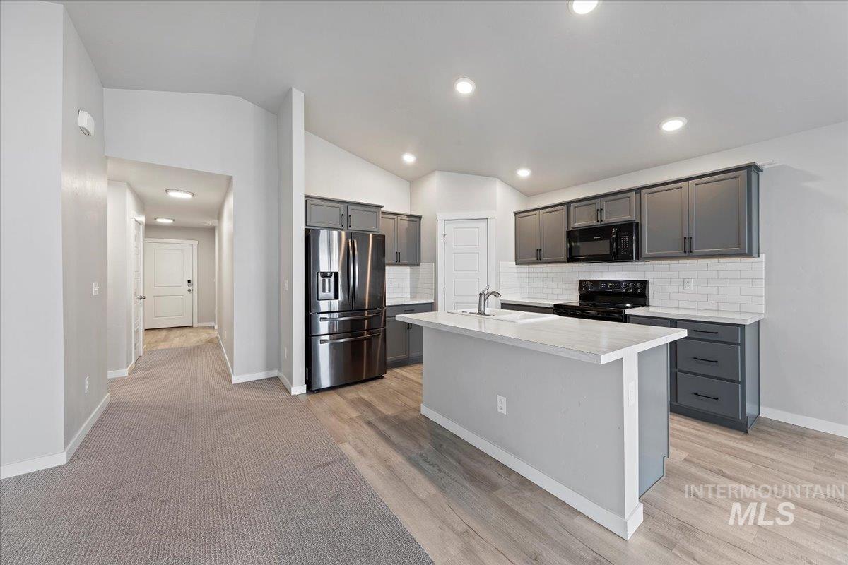 Kitchen featuring light countertops, black appliances, decorative backsplash, lofted ceiling, and a center island with sink