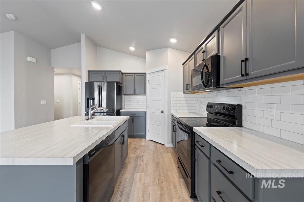 Kitchen featuring gray cabinetry, black appliances, light countertops, a kitchen island with sink, and lofted ceiling