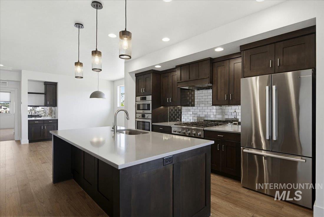 Kitchen featuring a central island with an undermount sink, pendant lighting, wood-finish flooring, dark cabinetry, and stainless steel appliances