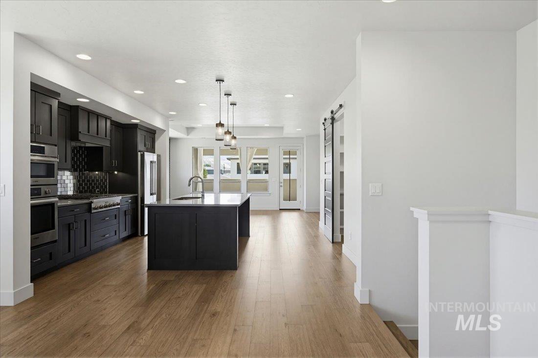 Modern kitchen featuring dark cabinetry, stainless steel appliances, a center island with sink, pendant lighting, and wood-finish flooring