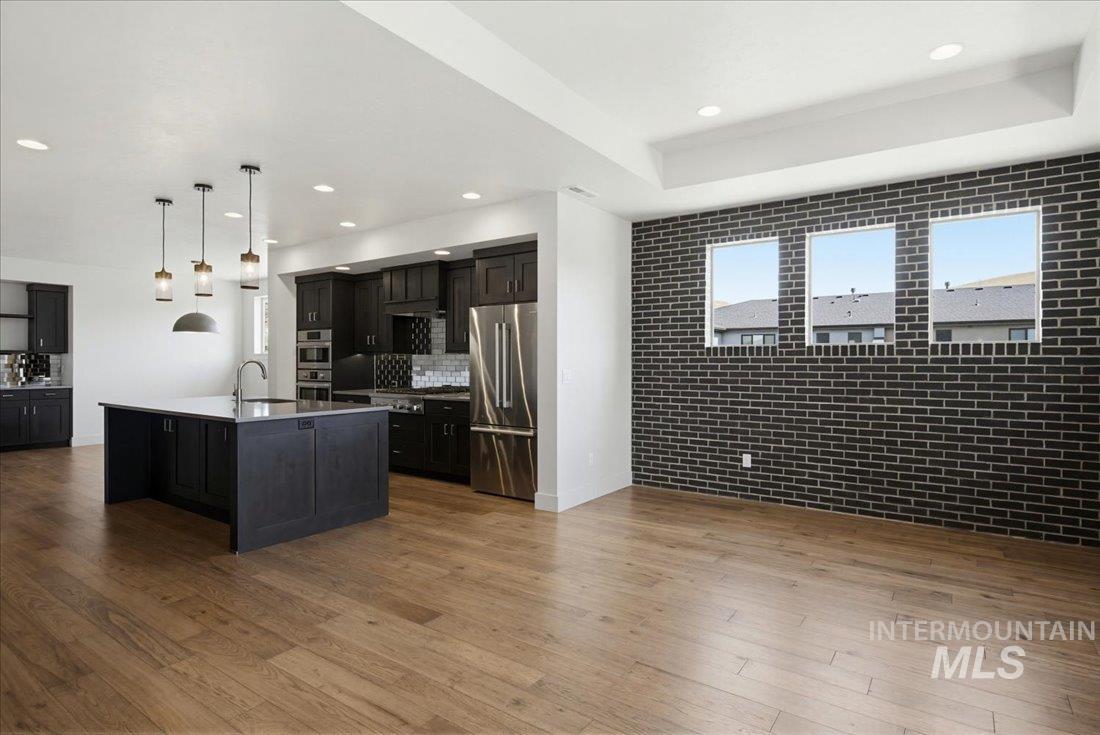 Open-concept living space featuring wood-finish flooring, a contemporary kitchen with dark cabinetry and stainless steel appliances, and a distinct black brick accent wall with three windows
