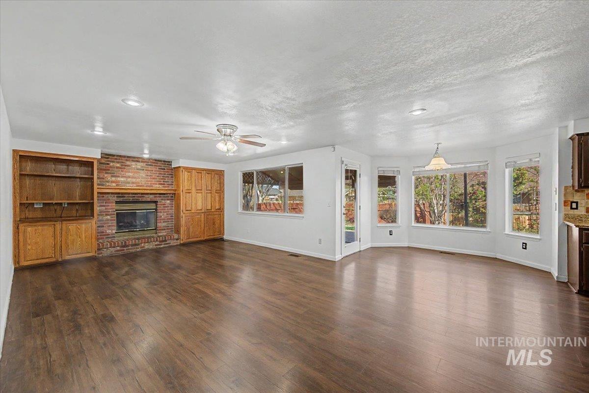 Unfurnished living room with dark wood-style flooring, a brick fireplace, ceiling fan, built in shelves, and a textured ceiling