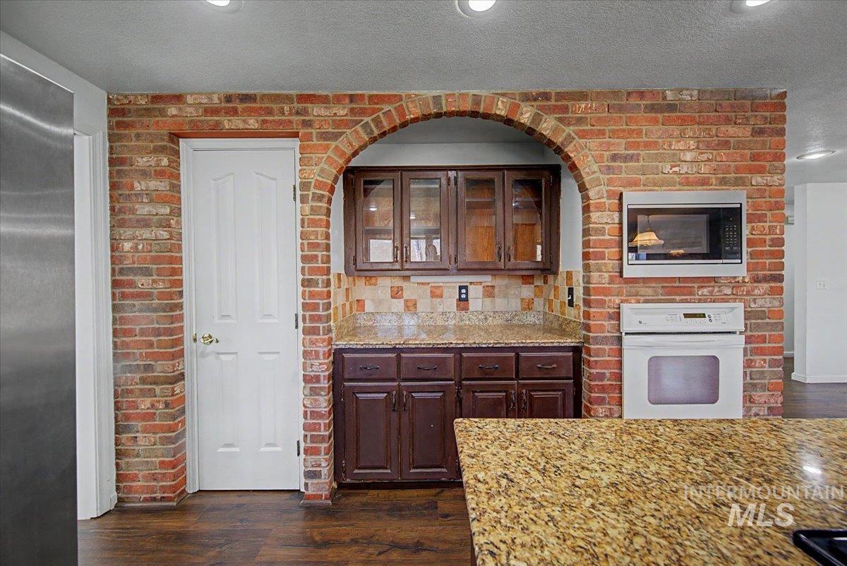 Kitchen with stainless steel appliances, glass insert cabinets, dark wood finished floors, brick wall, and light stone countertops