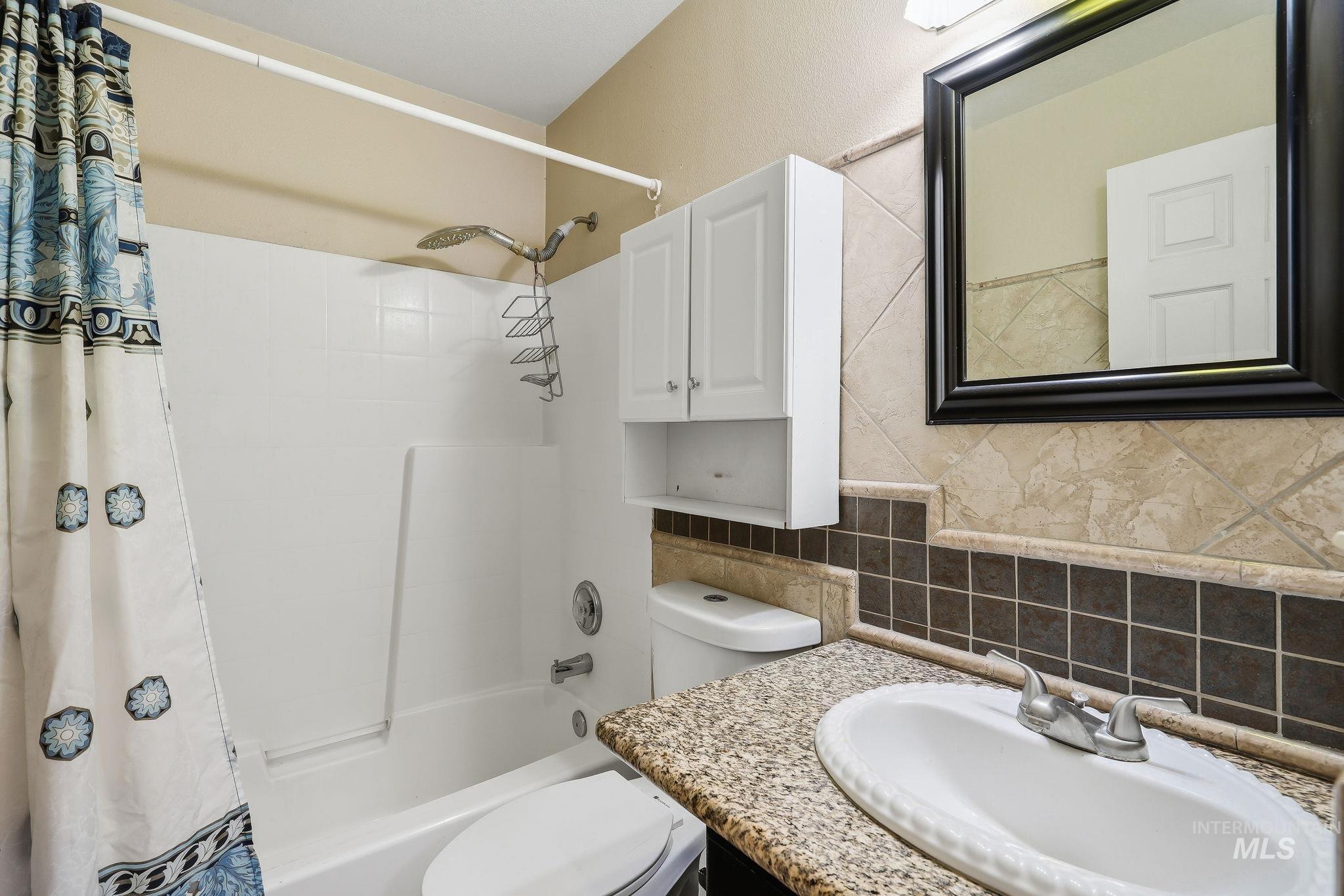 Bathroom featuring a white bathtub and shower combination, a granite-finish countertop with an integrated sink, a dark framed mirror, a wall-mounted storage cabinet, and tiled wall accents