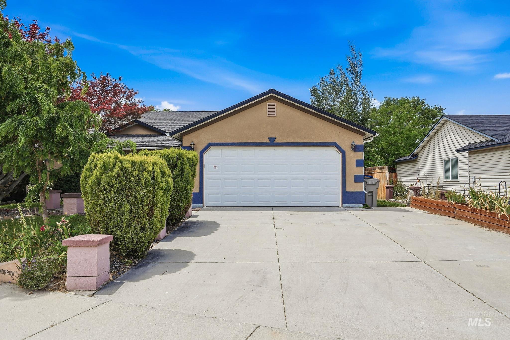 Exterior stucco facade with a two-car garage, concrete driveway, lush green shrubs, and mature trees