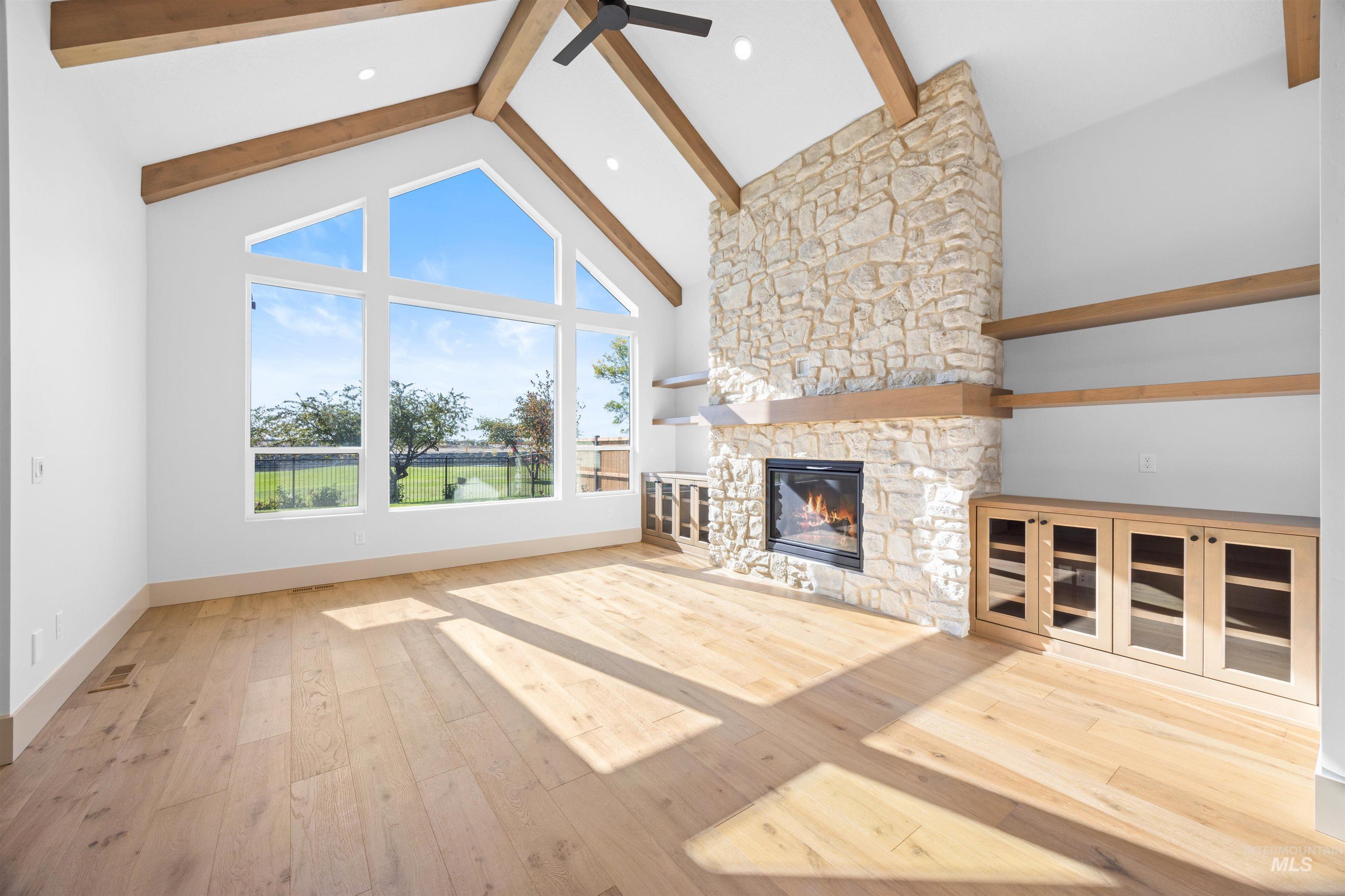 Unfurnished living room with beamed ceiling, high vaulted ceiling, light wood-style floors, a stone fireplace, and a ceiling fan