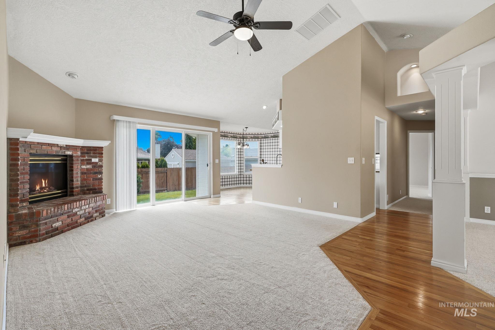 Spacious living area featuring a brick fireplace with a white mantel, sliding glass doors to the backyard, light-colored carpeting, a ceiling fan, and hardwood flooring at the entry
