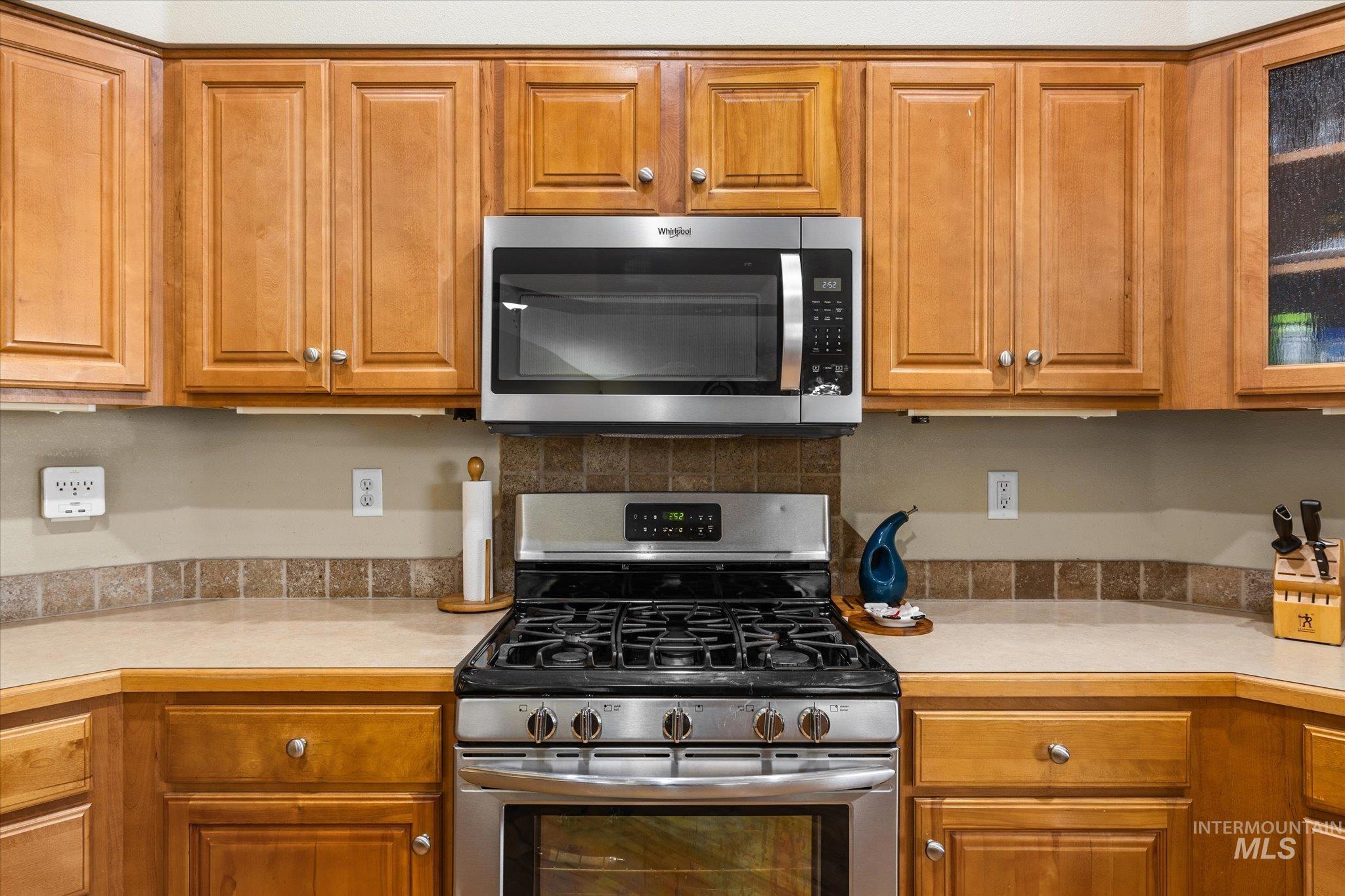 Kitchen featuring appliances with stainless steel finishes, light countertops, brown cabinets, glass insert cabinets, and decorative backsplash