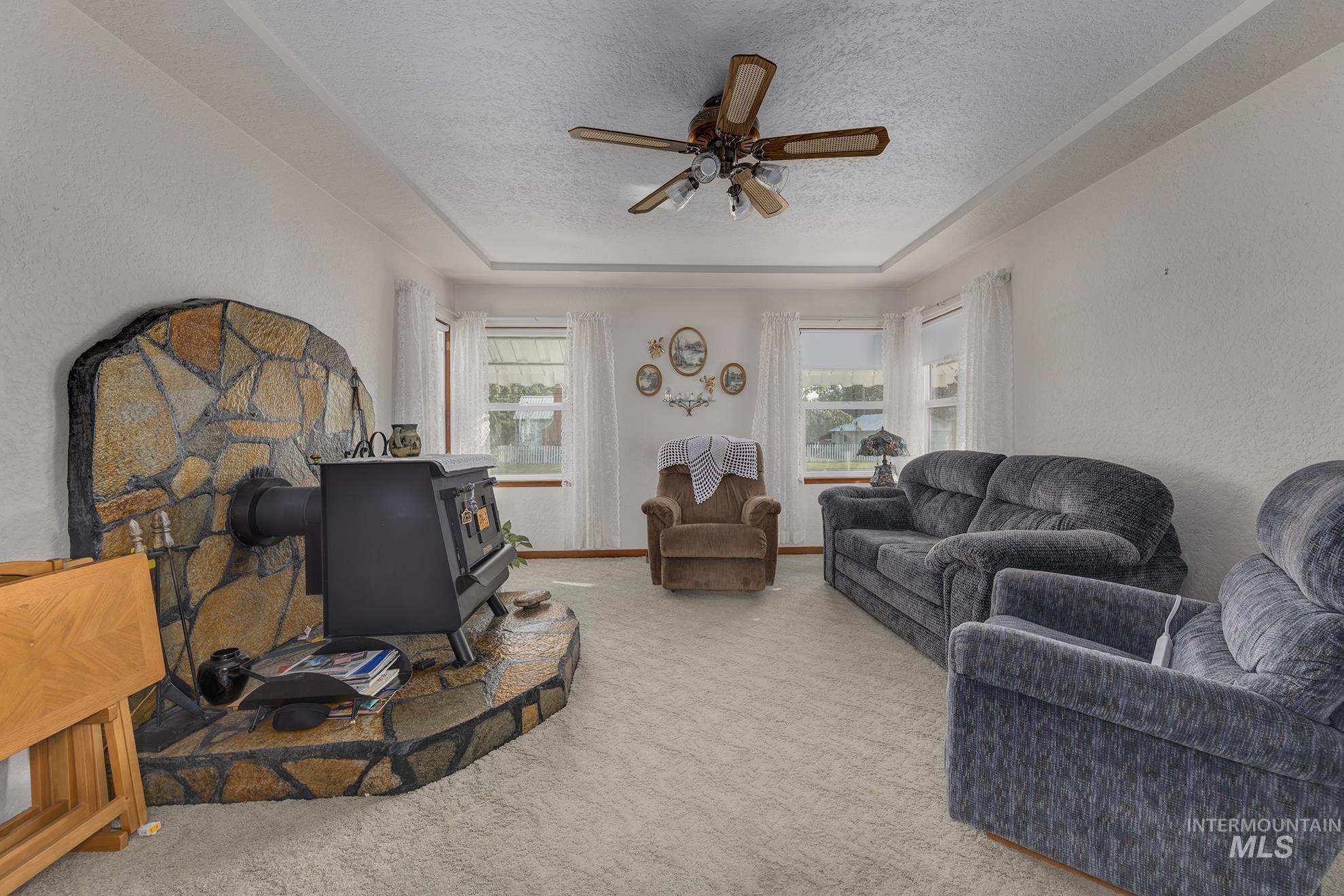 Living area with a wood stove, a textured ceiling, carpet floors, ceiling fan, and a tray ceiling