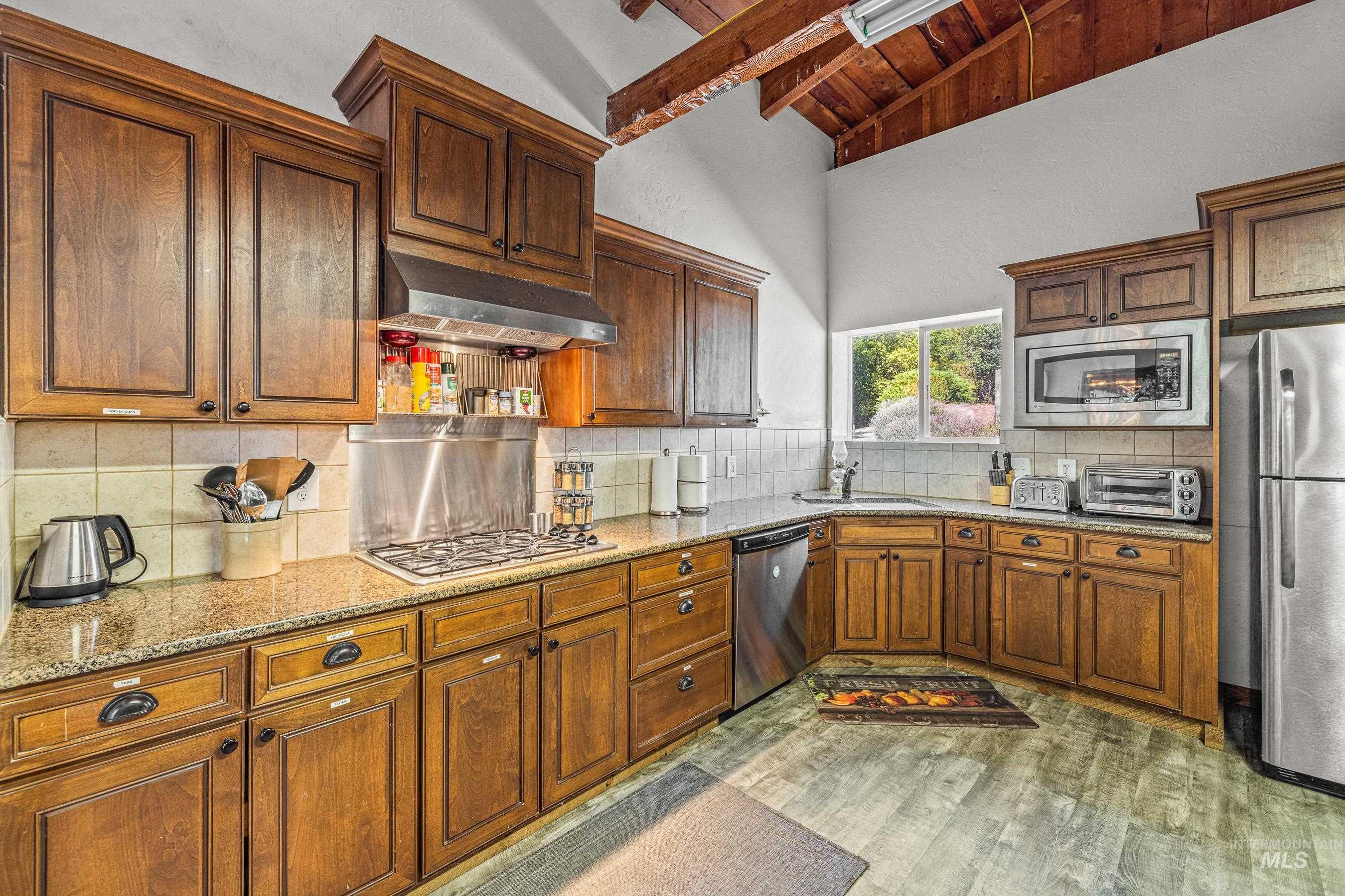 Kitchen with tasteful backsplash, stainless steel appliances, light stone counters, light wood-type flooring, and a wood ceiling with exposed beams