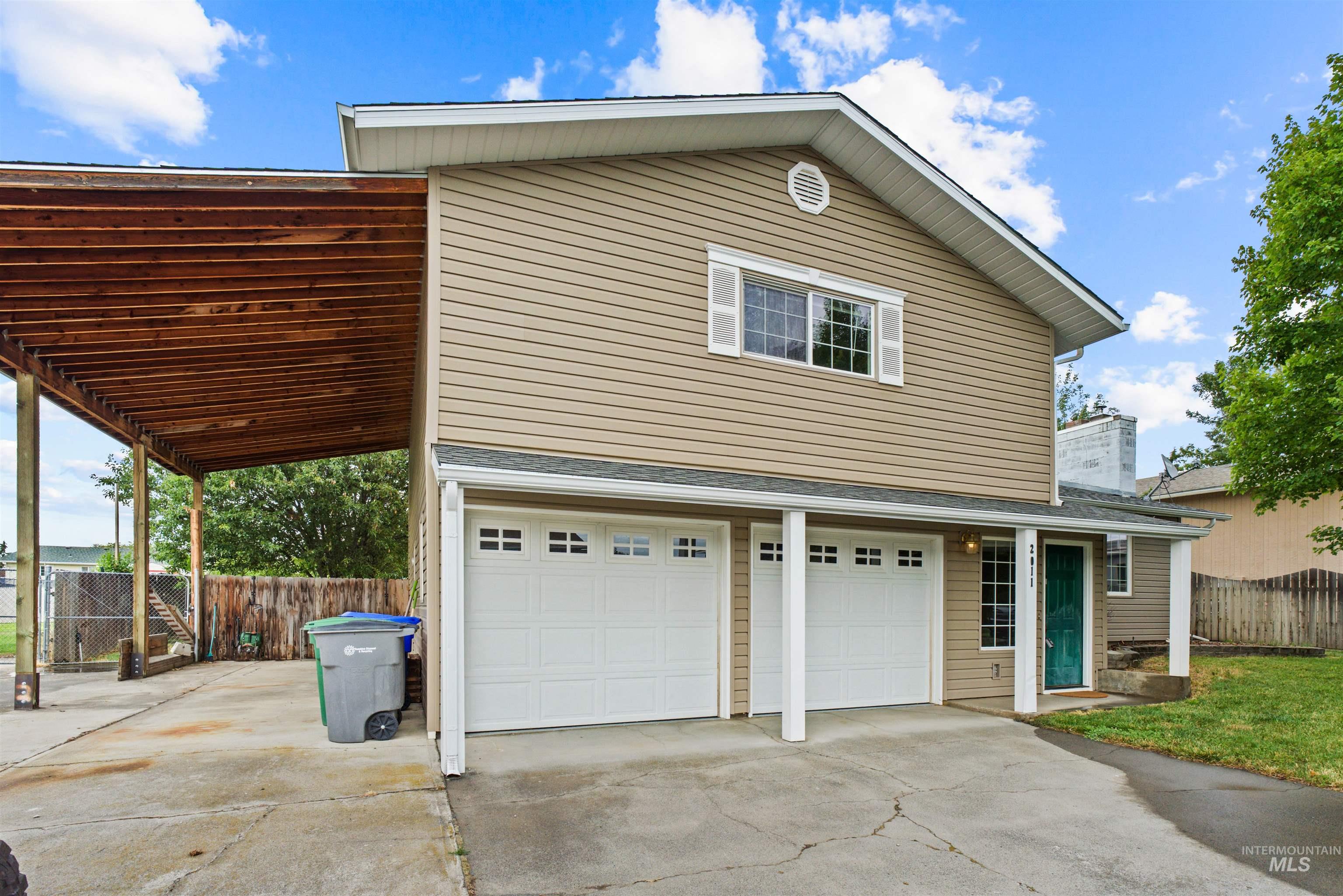 Two-story residence featuring tan siding, a two-car garage with white doors, and a covered carport with exposed wood beams