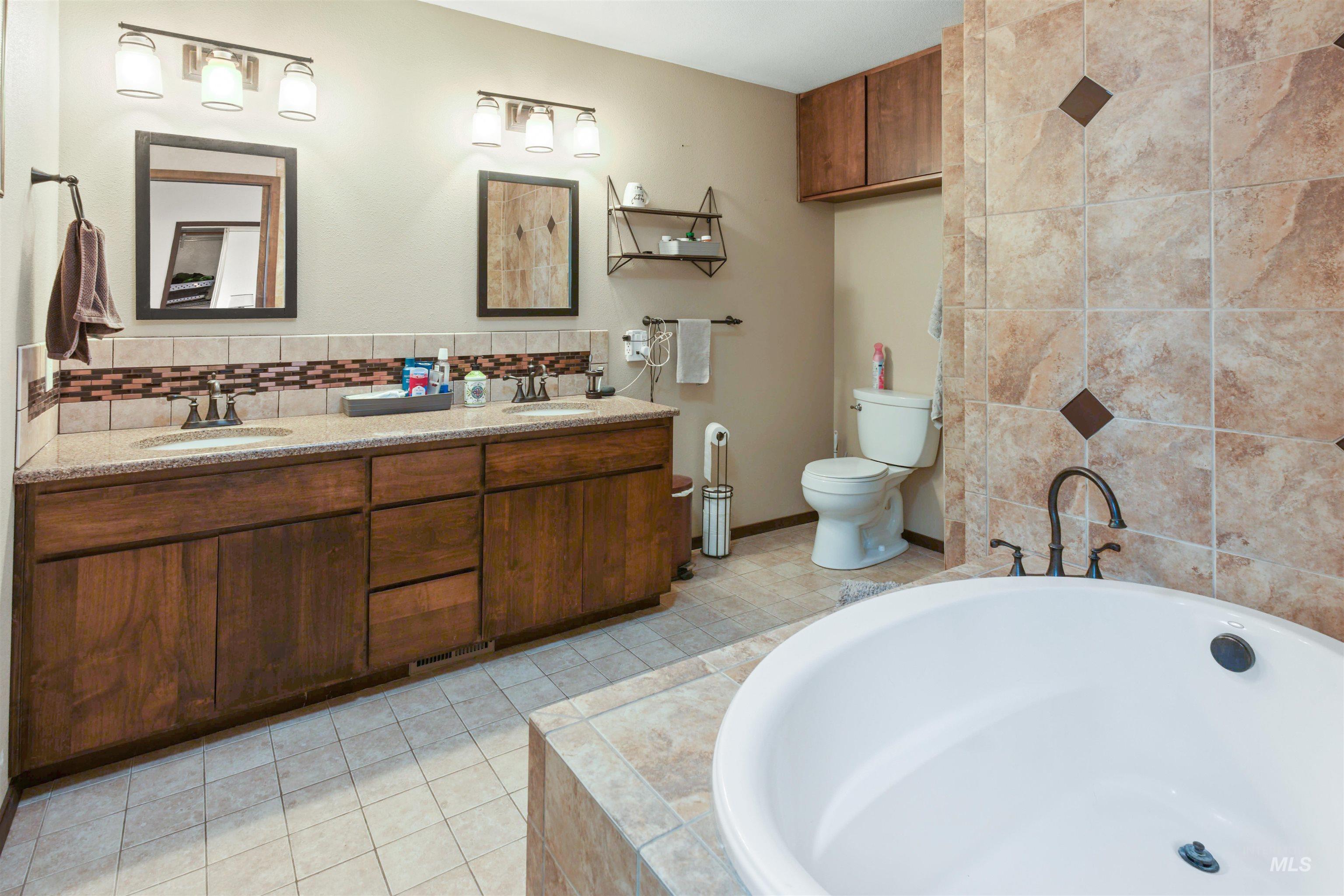 Bathroom featuring a dual vanity with wood cabinetry and light-toned countertops, a built-in soaking tub, and tiled floors