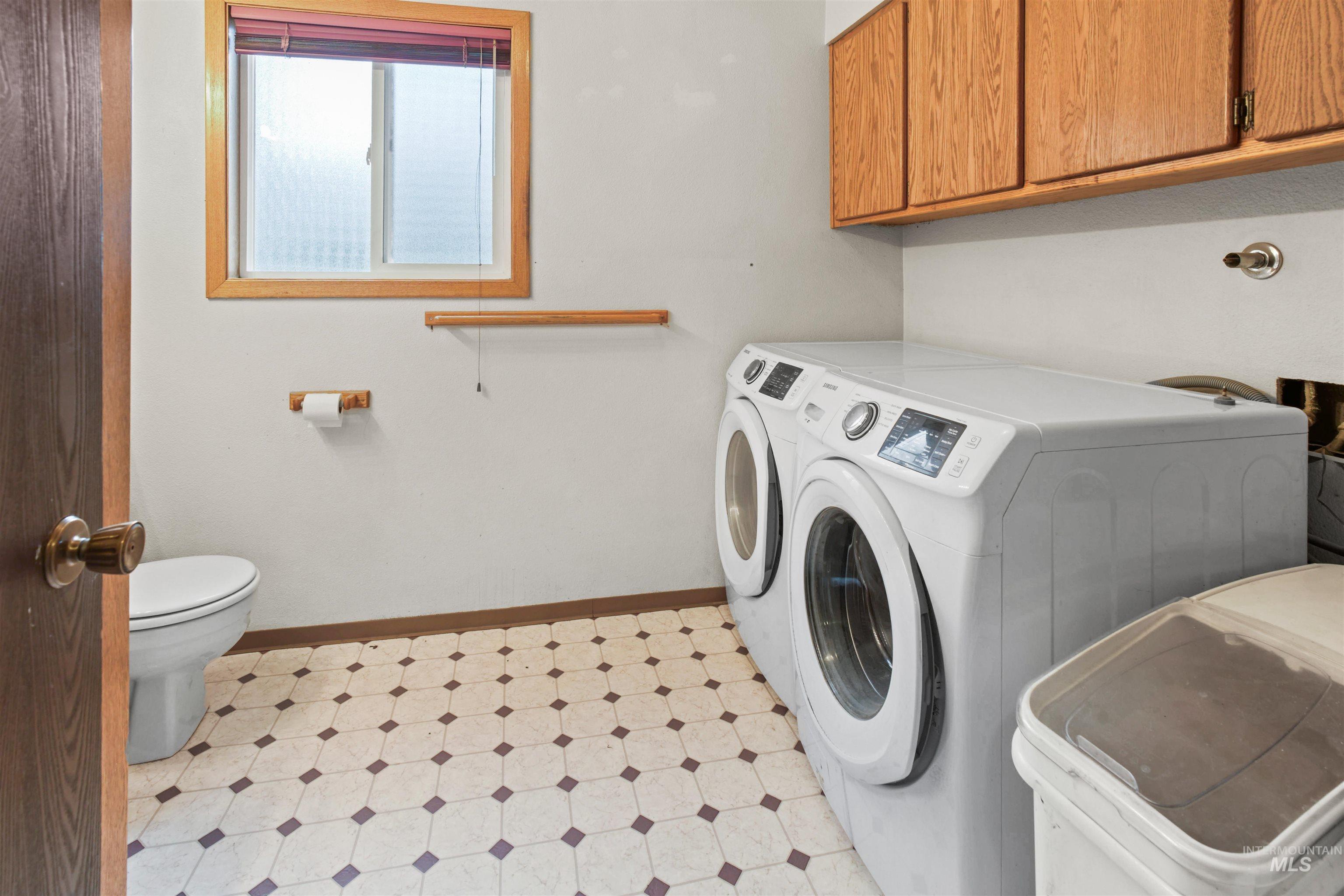Laundry area featuring front-loading washer and dryer, wood-finish cabinetry, a toilet, a window with wood trim, and patterned flooring