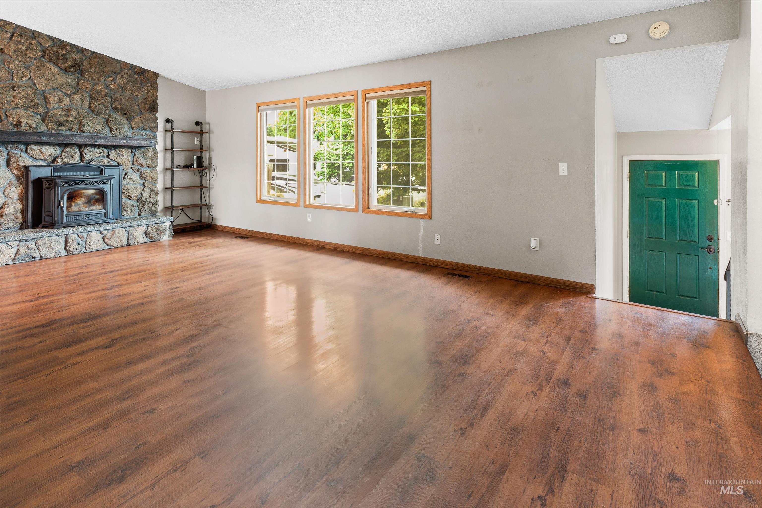 Spacious room featuring wood-finish flooring, a prominent stone fireplace with an insert, three large windows with wood trim, and a deep green entry door