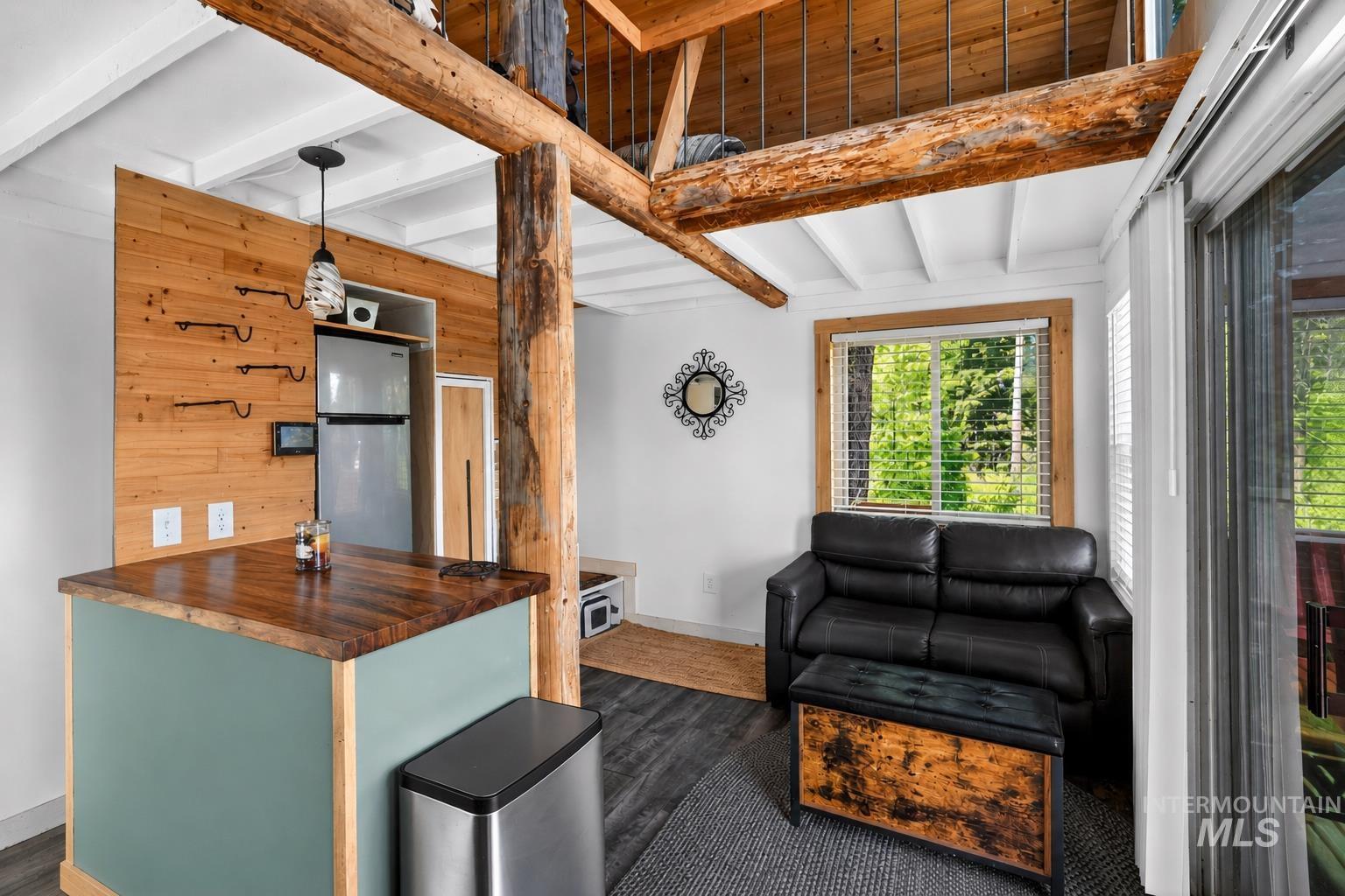 Open-concept living space featuring wood-finish flooring, exposed timber beams, a white-painted wood ceiling with exposed joists, a kitchen island with a wood countertop, and a window with natural scenery