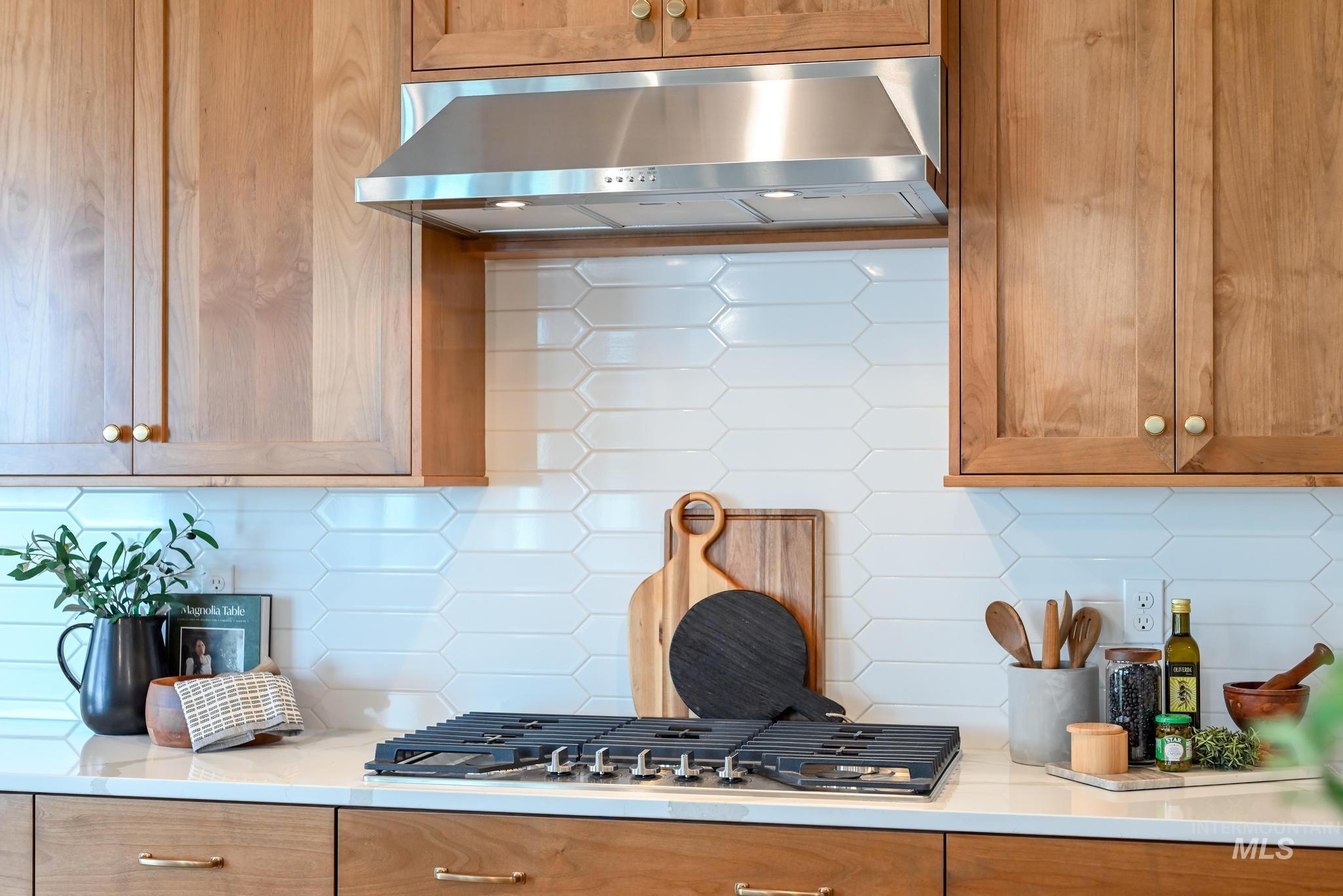 Kitchen with under cabinet range hood, backsplash, light stone counters, and brown cabinetry