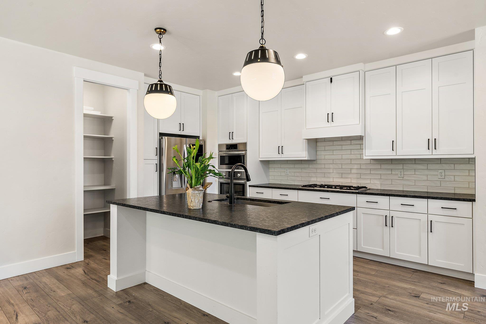 Kitchen featuring an island with sink, backsplash, hanging light fixtures, white cabinets, and dark wood-style flooring
